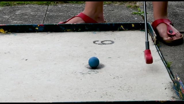 Eine Frau in roten Sandalen auf Minigolfplatz schl&auml;gt den Minigolfball. Der Ball rollt &uuml;ber die Bahn. Die Frau geht weg