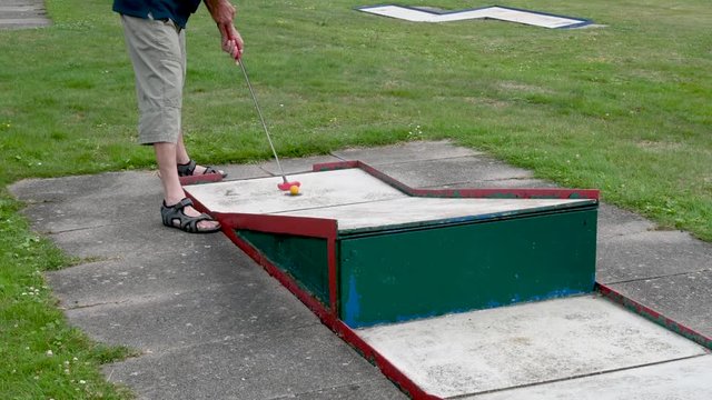 Mann in kurzer Hose auf Minigolfplatz schl&auml;gt den Minigolfball. Der Ball springt &uuml;ber die Bahn. Der Mann geht weg
