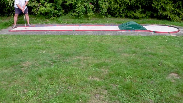 Mann in kurzer Hose auf Minigolfplatz schl&auml;gt den Minigolfball. Der Ball rollt &uuml;ber die Bahn und f&auml;llt nach einem zweiten Schlag mit dem Minigolfschl&auml;ger ins Loch.