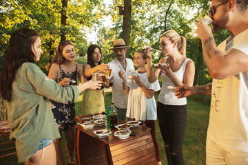 Group of happy friends eating and drinking beers at barbecue dinner on sunset time. Having meal together outdoor in a forest glade. Celebrating and relaxing. Summer lifestyle, food, friendship concept