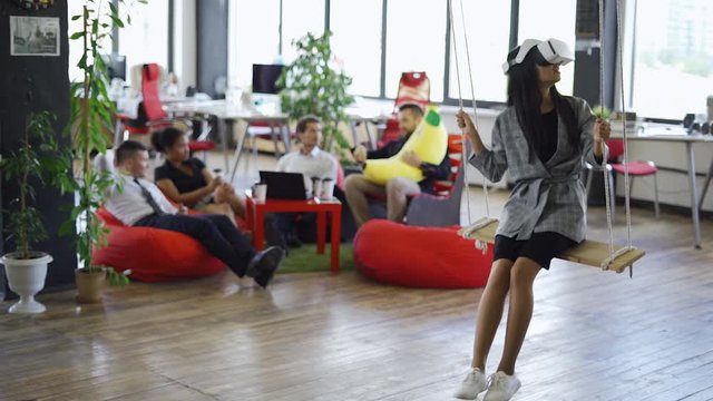 Young asian girl in futuristic white virtual reality glasses riding on a swing, modern office and workers on a blurry backround.