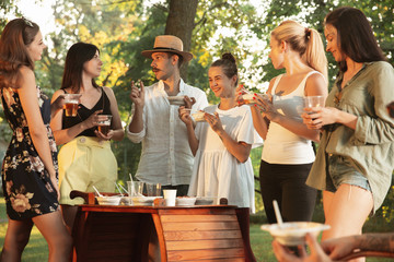 Group of happy friends eating and drinking beers at barbecue dinner on sunset time. Having meal together outdoor in a forest glade. Celebrating and relaxing. Summer lifestyle, food, friendship concept
