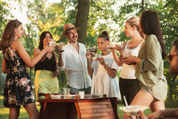 Group of happy friends eating and drinking beers at barbecue dinner on sunset time. Having meal together outdoor in a forest glade. Celebrating and relaxing. Summer lifestyle, food, friendship concept