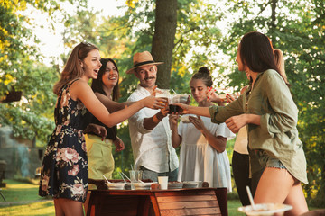 Group of happy friends eating and drinking beers at barbecue dinner on sunset time. Having meal together outdoor in a forest glade. Celebrating and relaxing. Summer lifestyle, food, friendship concept