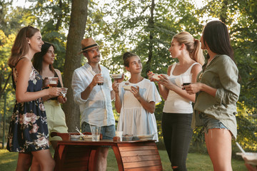 Group of happy friends eating and drinking beers at barbecue dinner on sunset time. Having meal together outdoor in a forest glade. Celebrating and relaxing. Summer lifestyle, food, friendship concept