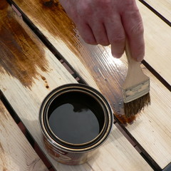 Hand holding a brush applying varnish paint on a wooden surface. Brush on wooden background. Board...