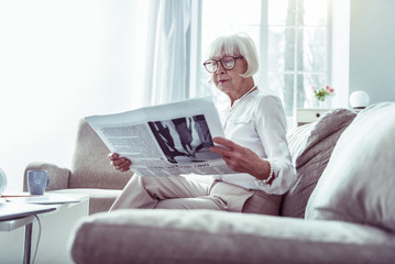 Beautiful elderly woman reading newspaper while waiting for husband at home