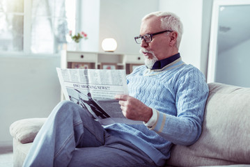 Bearded retired man wearing glasses sitting on sofa and reading newspaper