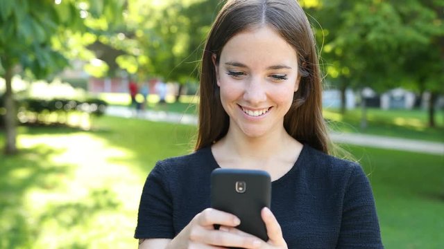 Front view of a happy woman walking towards camera in a park using smart phone