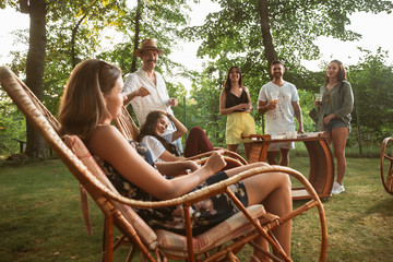 Group of happy friends eating and drinking beers at barbecue dinner on sunset time. Having meal together outdoor in a forest glade. Celebrating and relaxing. Summer lifestyle, food, friendship concept