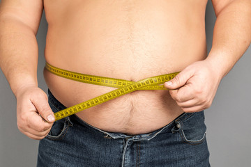 A man measures his fat belly with a measuring tape. on a gray background.