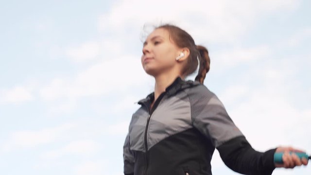 Tilt down of young Caucasian woman wearing sportswear listening to music using wireless headphones while skipping with jump rope on wooden embankment outside