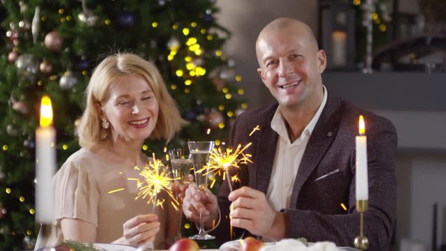 Waist-up shot of middle-aged Caucasian husband in suit and wife in party dress sitting at romantic dinner near Christmas tree, holding sparklers and champagne, posing for camera with happy smiles