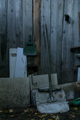 A homemade shovel rests on the wall of a wooden fence with plastic bins, a canister is lying nearby and a fan is hanging for barbecue