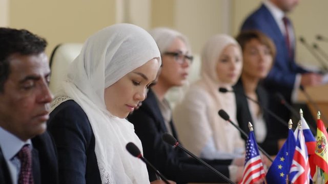 Muslim woman wearing hijab sitting at table with multiethnic group of politicians and giving a public talk during press conference