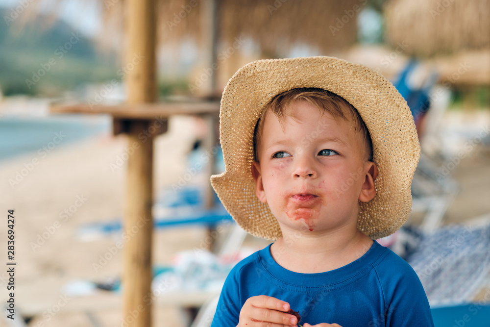 Wall mural Two year old toddler boy on beach eating fruit. Dirty messy face of happy kid.