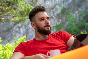 Stylish young man in a red with a cup of coffee. Young bearded hipster relaxing in the nature, drinking hot coffee.