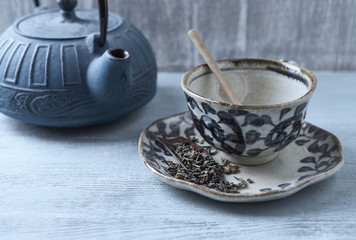 Empty Ceramic Tea Cup and Green Tea Leaves. Iron Kettle in Background. Symbolic image. Asian culture. Traditional Japanese Ceramic. Rustic wooden background. Close up. 