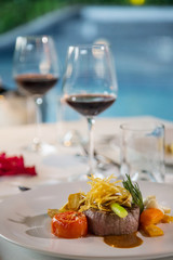 Beefsteak with steamed vegetables and glasses of red wine served on the table, Romantic dinner next to the pool, outdoor summer dining concept