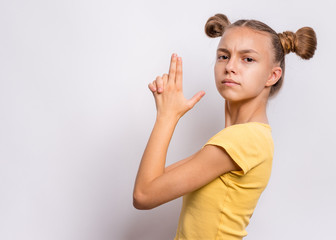 Fototapeta premium Portrait of funny teen girl doing gun gesture getting ready to shoot. Caucasian young teenager showing raised gun gesture on gray background. Child holding symbolic gun. Emotions and signs.