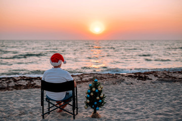 A guy in a santa hat on the beach with a Christmas tree at sunset.