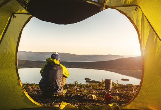 View from inside a tent. The traveler sits in front of the tent on the mountain and lakes at light of sunset background. Kungsleden. 