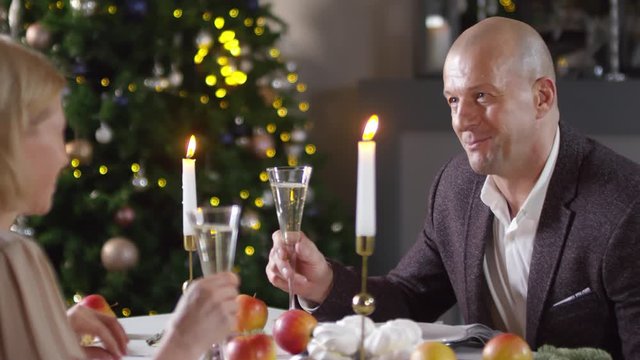Chest-up shot of handsome 40-something Caucasian man in smart blazer, with shaved head, having romantic dinner with girlfriend on New Year&rsquo;s Eve, holding up glass of champagne and saying long toast