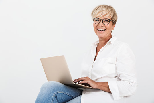 Image of beautiful adult woman wearing eyeglasses laughing while sitting with laptop computer