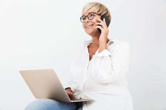 Image of gorgeous adult woman wearing eyeglasses talking on smartphone while sitting with laptop computer