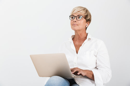 Image of middle-aged woman wearing eyeglasses and earpods sitting with laptop computer