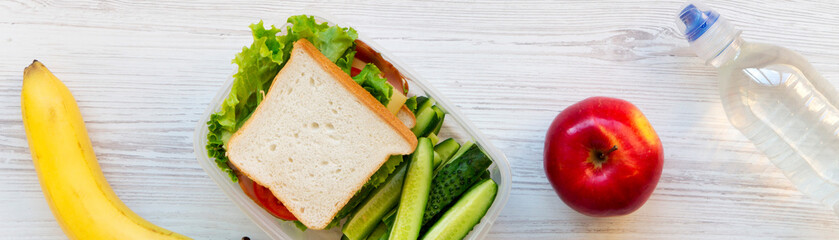 School lunch box with sandwich, fruits and bottle of water on a white wooden surface, top view. From above, overhead.