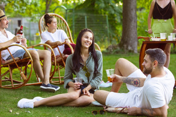 Group of happy friends eating and drinking beers at barbecue dinner on sunset time. Having meal together outdoor in a forest glade. Celebrating and relaxing. Summer lifestyle, food, friendship concept