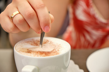 Closeup of a woman's hand with an golden wedding ring stirring her cappuccino in a white cup