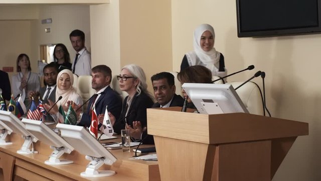 Cheerful muslim woman in hijab walking to rostrum while multiethnic participants clapping hands and then start speaking to audience during political press conference