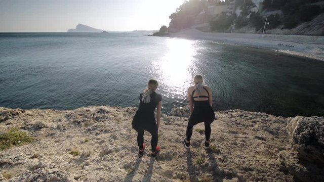 Two young women doing exercises by the sea. Slim attractive caucasian girls practicing yoga doing breathing exercise on small bay background. Back view.