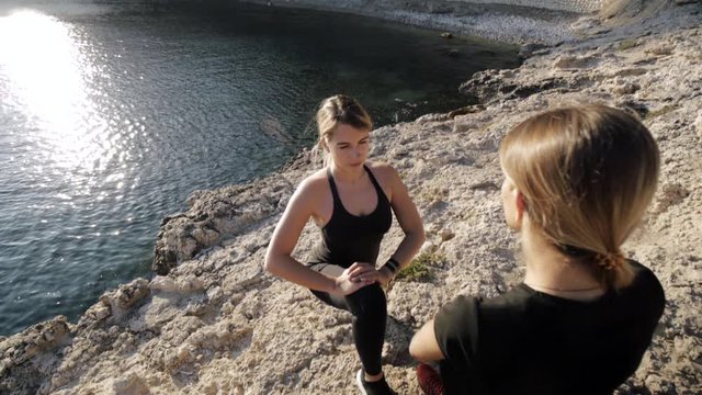 Two young women doing exercises by the sea. Slim attractive caucasian girls practicing yoga doing stretching exercise on rocky beach. Pull out shot. Slow motion.