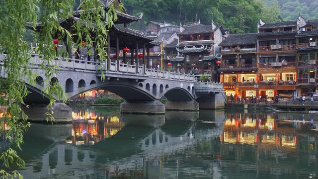 Fenghuang Bridge across Tuojiang River in Fenghuang, China before dusk. Lights from local restaurants are reflecting on the river's surface. UHD