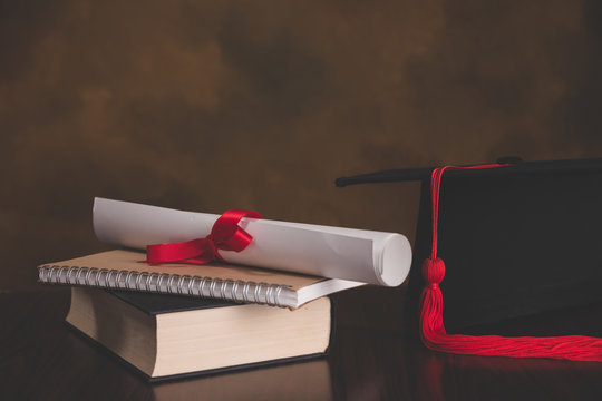 A mortarboard and graduation scroll, tied with red ribbon, on a stack of old battered book with empty space to the left. Slightly undersaturated with vignette for vintage effect.