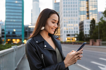 Fototapeta premium Smiling happy young woman walking outdoors by street in city using mobile phone.