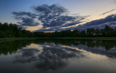 Beautiful evening landscape by the forest lake. Trees, sky and clouds are reflected in the water
