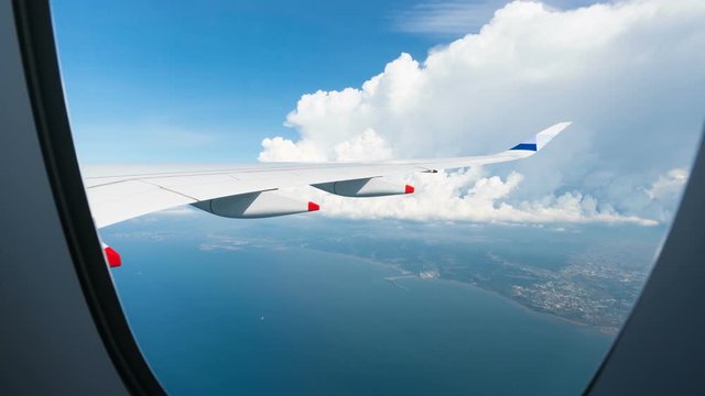 Zooming in timelapse view through plane illuminator of passenger jet wing flying above great blue sea, view of light white clouds and bright blue sky