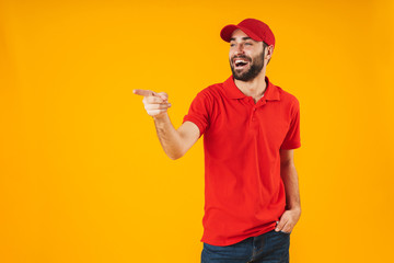 Portrait of positive delivery man in red t-shirt and cap smiling and pointing finger at copyspace