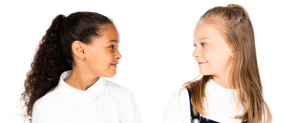 panoramic shot of two smiling multicultural schoolgirls looking at each other isolated on white