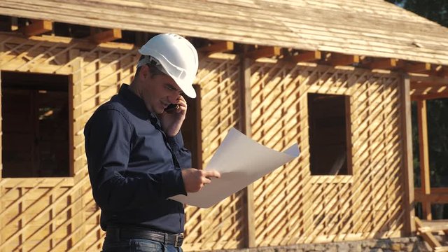 concept building constructing architect slow motion video. man builder in a helmet stands at a construction lifestyle holding a scheme house plan. site near a wooden frame house under construction