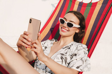 Cheerful young pretty woman at the beach posing on a hammock take a selfie by mobile phone.