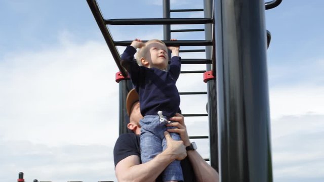 Medium shot of happy young Caucasian father hanging out together with 3-year-old blond toddler son at outdoor workout gym, holding boy up and helping him move across horizontal hand ladder