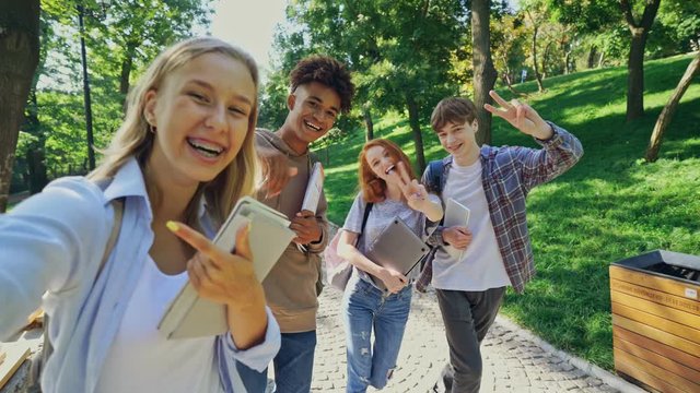 Group of cheerful multiethnic students having fun together and making selfie while walking outdoors in park