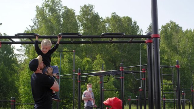Full shot of happy Caucasian toddler hanging on monkey bar and pulling up on grip at outdoor gym, dad holding and lifting him up, and young father with baby daughter and pram walking in background