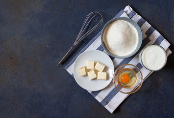 Ingredients for crepes and pancakes on a dark background. Flour, egg, milk, butter, whisk. Top view. Space for text