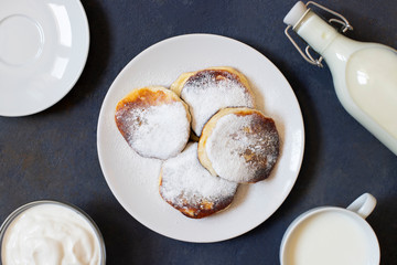 Cottage cheese pancakes. Cheesecakes. Syrniki or sirniki. Healthy breakfast. White dishes. Bottle of milk. A cup of milk. Yogurt or sour cream. Dark background. Top view. Space for text